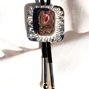 May include: A bolo tie with a rectangular, multi-colored stone set in a silver-toned, square frame with a wavy border. The black cord has silver tips with small, round accents.