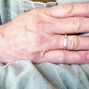 May include: A close-up of a person's hand wearing a silver band ring. The ring is simple and has a slightly textured surface.