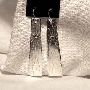 May include: A pair of silver-toned earrings. Each earring is a long, rectangular shape with a sunburst design at the top and etched lines. The earrings hang from a simple hook. The background is a white fabric.