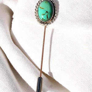 May include: A silver-toned lapel pin with an oval turquoise stone. The stone is set in a twisted rope-like border. The pin has a long, thin, copper-colored stem and a silver-toned, cylindrical end.