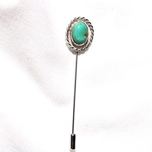 May include: A silver-toned stick pin with an oval turquoise stone. The stone is set in a decorative frame with a twisted rope design. The pin is a classic accessory.