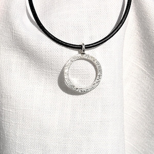 May include: A silver pendant necklace featuring a textured, open circle design. The pendant hangs from a black cord. The necklace is displayed against a white fabric background, creating a contrast that highlights the jewelry.