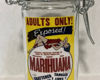 May include: A glass jar with a metal latch closure. The jar has a yellow label with the text "ADULTS ONLY! Exposed!" and "MARIHUANA" in red letters. The label also includes the text "DIVULGING HERETOFORE UNHEARD OF ORGIES OF YOUTH'S DISSIPATION SENSATIONAL! DARING! BOLD!" and "SHATTERED HOPES! TANGLED LIVES!"