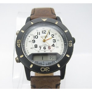 May include: A black and gold Guess watch with a brown leather band. The watch has a digital display and an analog face with white hands and black numbers. The watch is water resistant and has a stop watch function.