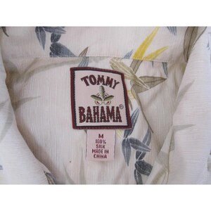 Może przedstawiać: Zbliżenie kremowej jedwabnej koszuli z kwiatowym wzorem. Koszula posiada metkę Tommy Bahama z napisem "M 100% Silk Made in China". Koszula ma kołnierzyk.
