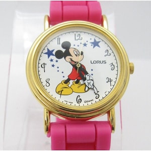 May include: A gold-tone Mickey Mouse watch with a white face and pink silicone band. The watch features a Mickey Mouse illustration with blue stars and the text "Lorus" and "©Disney".