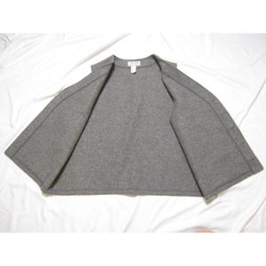 Puede incluir: Una chaqueta gris de estilo capa con frente abierto. La chaqueta est&aacute; hecha de un material suave similar al fieltro y presenta un dise&ntilde;o simple y minimalista. La prenda tiene una peque&ntilde;a etiqueta en el escote y un cierre de bot&oacute;n en el lado derecho.