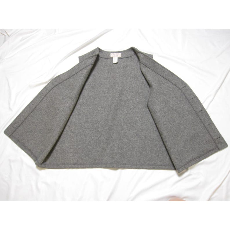 Puede incluir: Una chaqueta gris de estilo capa con frente abierto. La chaqueta est&aacute; hecha de un material suave similar al fieltro y presenta un dise&ntilde;o simple y minimalista. La prenda tiene una peque&ntilde;a etiqueta en el escote y un cierre de bot&oacute;n en el lado derecho.