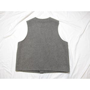 Puede incluir: Un chaleco de lana gris con cuello redondo y cierre de botones. La prenda sin mangas es de color liso y parece estar hecha de un material suave y texturizado. El chaleco se muestra sobre un fondo blanco.