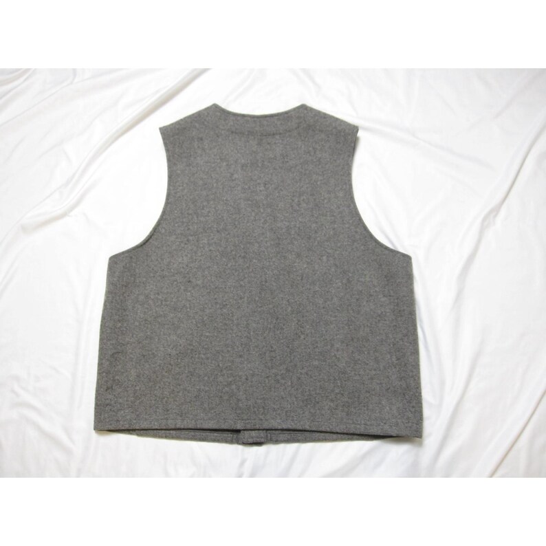 Puede incluir: Un chaleco de lana gris con cuello redondo y cierre de botones. La prenda sin mangas es de color liso y parece estar hecha de un material suave y texturizado. El chaleco se muestra sobre un fondo blanco.