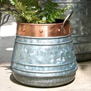 May include: A galvanised metal planter with a copper top rim and a handle. The planter has a distressed finish and is filled with green ferns.