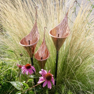 May include: Three copper sculptures of stylised calla lilies with black metal stems, set in a garden with pink coneflowers and tall, feathery grasses.