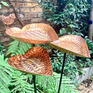 May include: Three copper heart-shaped bird feeders with a hammered texture, mounted on black metal poles, in a garden setting.