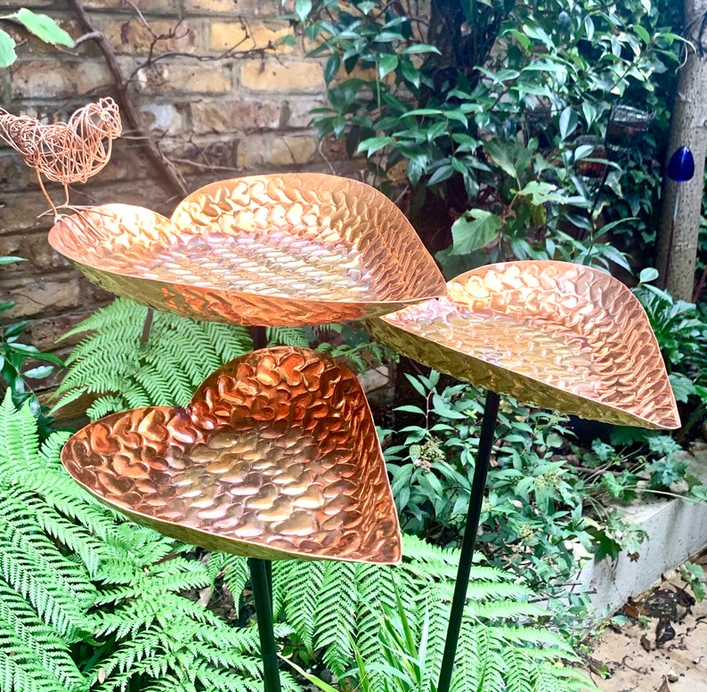 May include: Three copper heart-shaped bird feeders with a hammered texture, mounted on black metal poles, in a garden setting.