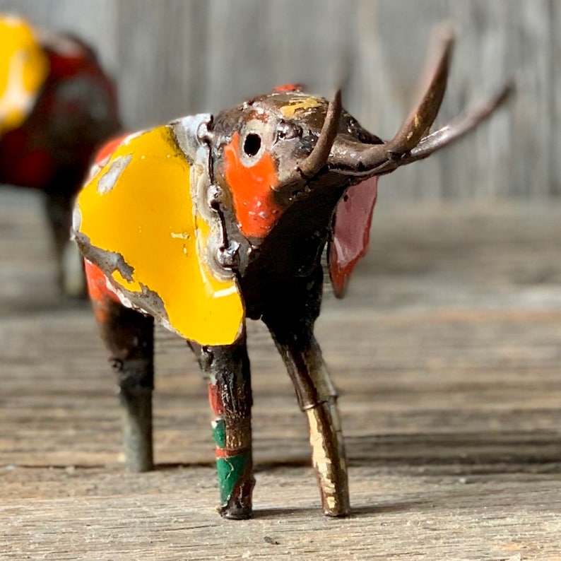 May include: A handcrafted metal elephant figurine with a distressed, colorful finish. The elephant features yellow, orange, red, and pink accents. The trunk is made of metal and the ears are pink. The figurine is on a wooden surface.