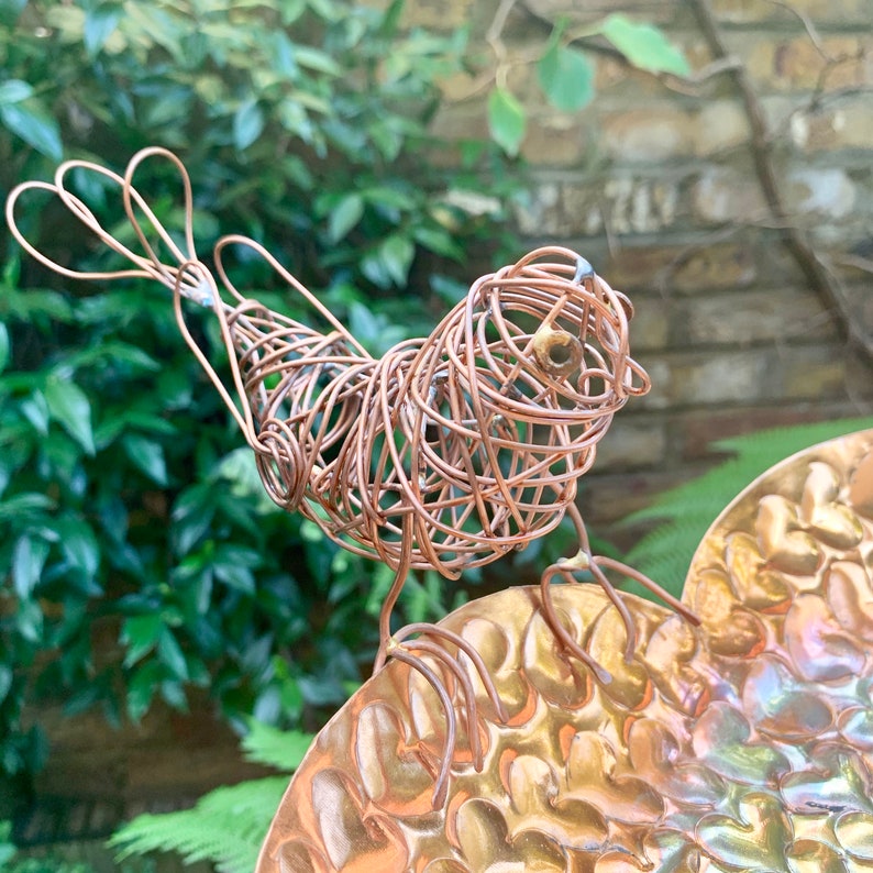May include: A copper wire bird sculpture perched on a hammered copper dish. The bird has a rounded body and a long tail. The dish has a textured surface.