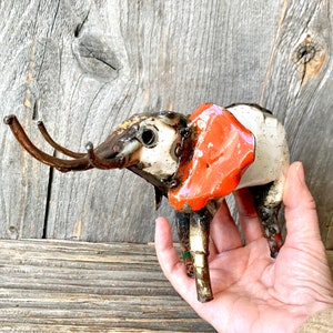 May include: A handcrafted metal elephant sculpture with a white body, orange ears, and brown tusks. The sculpture has a rustic, industrial aesthetic, with visible welds and a weathered appearance. The elephant is held in a hand against a wooden background.