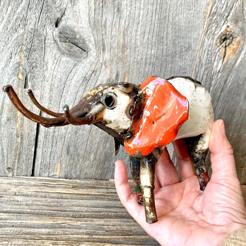 May include: A handcrafted metal elephant sculpture with a white body, orange ears, and brown tusks. The sculpture has a rustic, industrial aesthetic, with visible welds and a weathered appearance. The elephant is held in a hand against a wooden background.