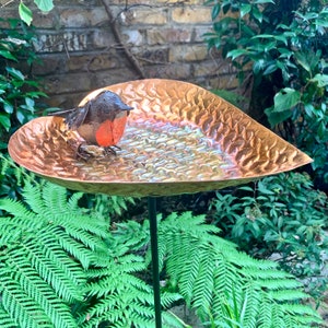 May include: A copper heart-shaped bird bath with a hammered texture and a small robin figurine perched on the edge.