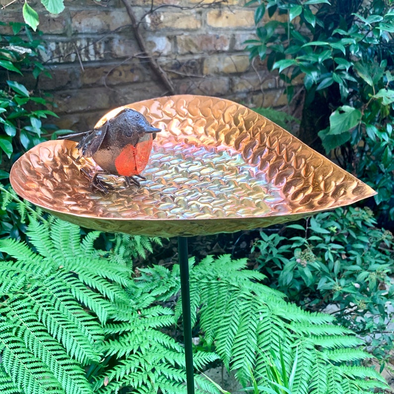 May include: A copper heart-shaped bird bath with a hammered texture and a small robin figurine perched on the edge.