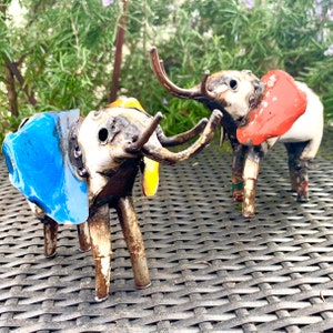 May include: Two handcrafted metal elephant sculptures. One elephant has blue ears, the other has red ears. Both have long, curved tusks and are painted with a mix of colors. The elephants are standing on a woven surface.