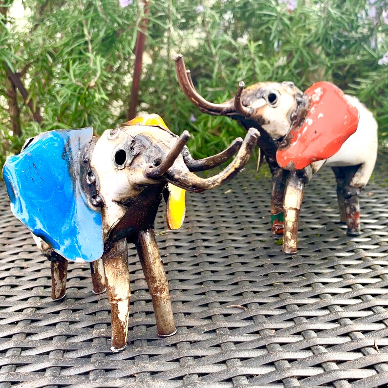 May include: Two handcrafted metal elephant sculptures. One elephant has blue ears, the other has red ears. Both have long, curved tusks and are painted with a mix of colors. The elephants are standing on a woven surface.