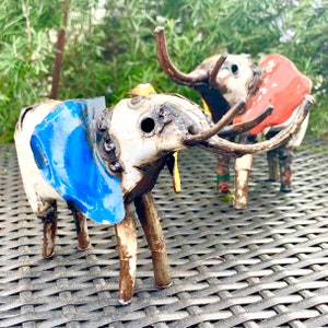 May include: Two handcrafted metal elephant sculptures. One elephant has blue ears, the other has red ears. Both have long tusks and are painted white with brown legs and trunks. The sculptures are on a dark gray woven surface.