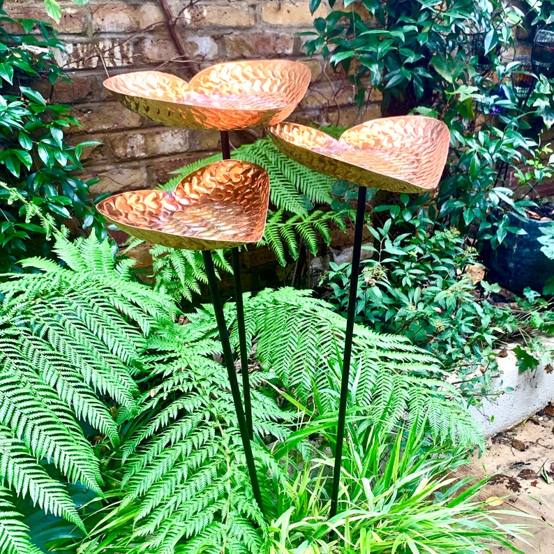 May include: Three copper heart-shaped bird feeders on black metal stakes, set in a lush green garden with ferns.