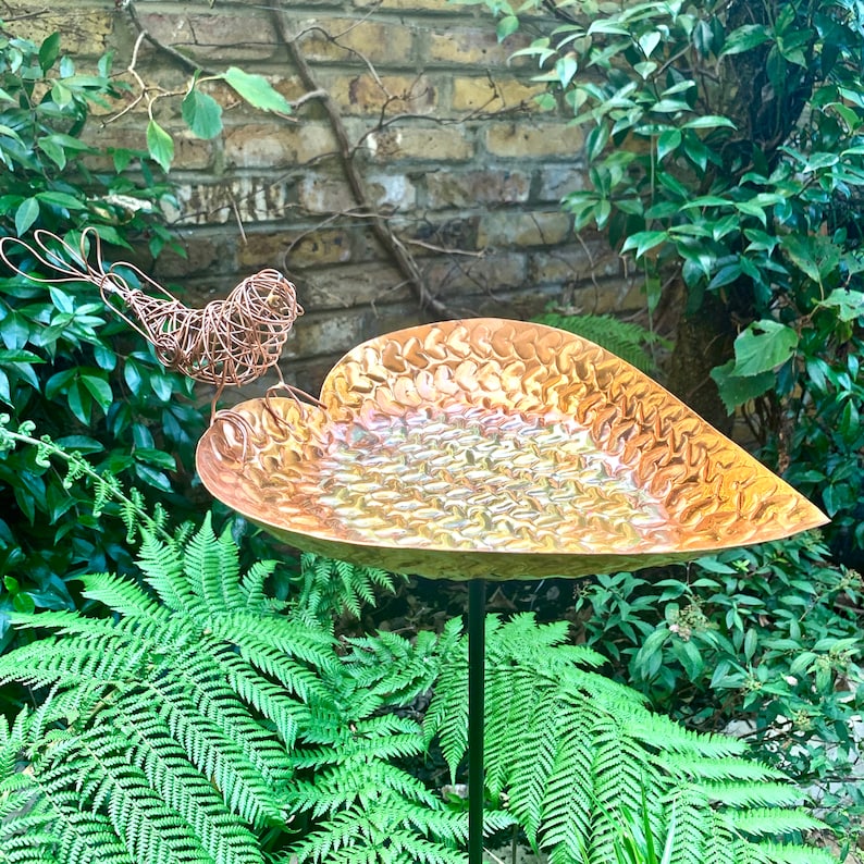 May include: A copper bird bath shaped like a leaf with a wire bird perched on the edge. The bird bath is on a black metal stand and surrounded by green ferns.