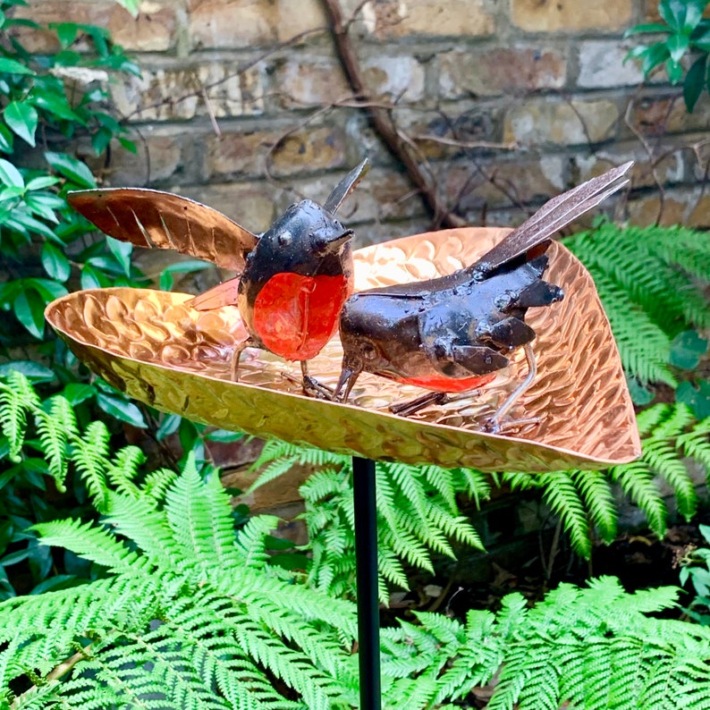 May include: Two black and red metal birds perched on a copper heart-shaped bird feeder. The feeder is attached to a black metal pole.