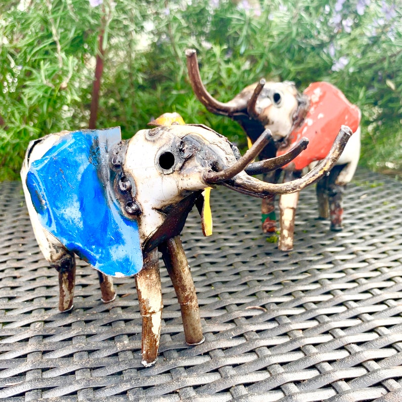 May include: Two handcrafted metal elephant sculptures. One elephant is painted white and blue, the other is white and red. Both have long tusks and are standing on a woven surface. The sculptures are outdoors with greenery in the background.