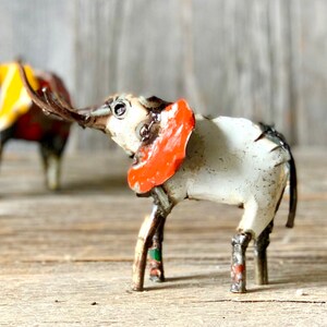 May include: A handcrafted metal elephant sculpture with a white body, orange ear accents, and brown legs. The elephant has a raised trunk and is set against a rustic wooden background. Another elephant is partially visible in the background.