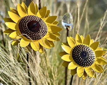 May include: Two yellow metal sunflower garden stakes with dark brown centers. The petals are curved and detailed, and the stakes are set against a backdrop of tall, dry grass. A clear glass orb is visible in the background.