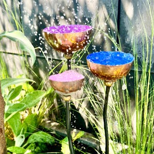 May include: A decorative outdoor water fountain with three copper-colored bowls, each holding a different color: purple, pink, and blue. The bowls are arranged at different heights and are connected by black stems. Water droplets are visible, suggesting the fountain is in operation.