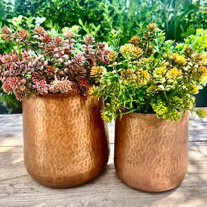 May include: Two copper-coloured, hammered metal planters, each filled with colourful faux plants. One planter holds reddish-pink flowers, the other holds yellow and green foliage. The planters sit on a wooden surface.
