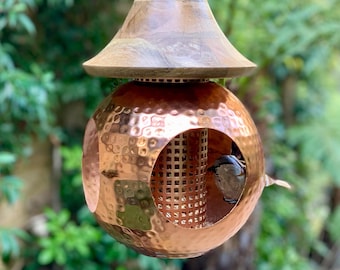 Puede incluir: Un comedero para pájaros de cobre con un acabado martillado y un techo de madera. Un pájaro está posado en el comedero, comiendo semillas.