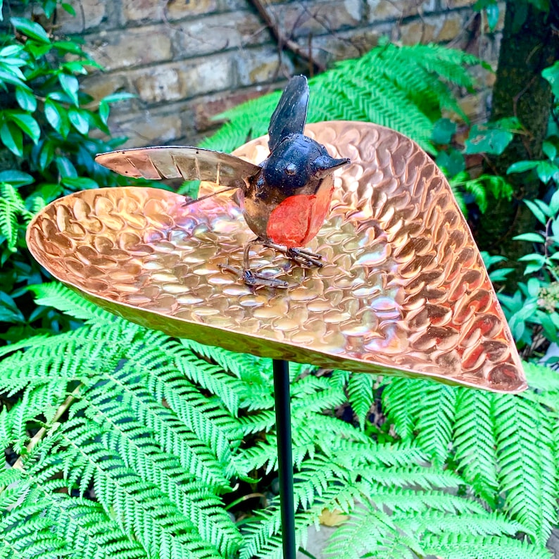 May include: A copper heart-shaped bird feeder with a black and red metal bird perched on the edge. The feeder is mounted on a black metal stake.
