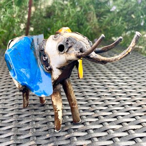 May include: A handcrafted metal elephant sculpture with a blue ear, white body, and brown tusks. The sculpture has a welded construction and a yellow accent. The elephant is standing on four metal legs.
