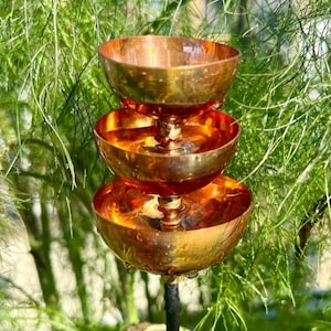 May include: A three-tiered copper bird bath with a shiny finish. The bird bath has three bowl-shaped tiers stacked on top of each other, supported by a black metal pole. The background is a blur of green foliage.