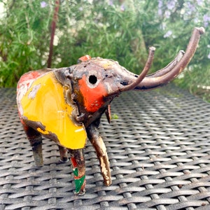 May include: A handcrafted metal elephant sculpture with a distressed, rustic appearance. The elephant features a yellow body with red and orange accents, and brown tusks. The sculpture is made from recycled metal.
