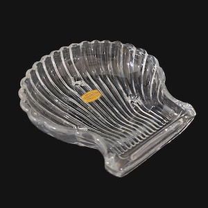 May include: Clear, lead crystal seashell-shaped dish with a ribbed design. The dish has a scalloped edge and a small, rectangular base. A gold sticker is affixed to the dish. The dish is on a black background.