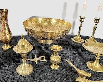 May include: A collection of antique brass decorative items, including a vase, bowl, candlesticks, and serving dishes. The items are ornate with etched designs and are arranged on a dark surface. Two candlesticks hold lit white candles.