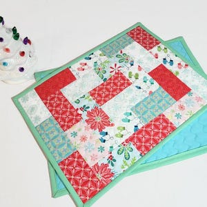 May include: Two quilted placemats with a Christmas theme. The placemats are made of red, white, blue, and green fabric with a variety of patterns. The placemats are edged in a light green fabric.