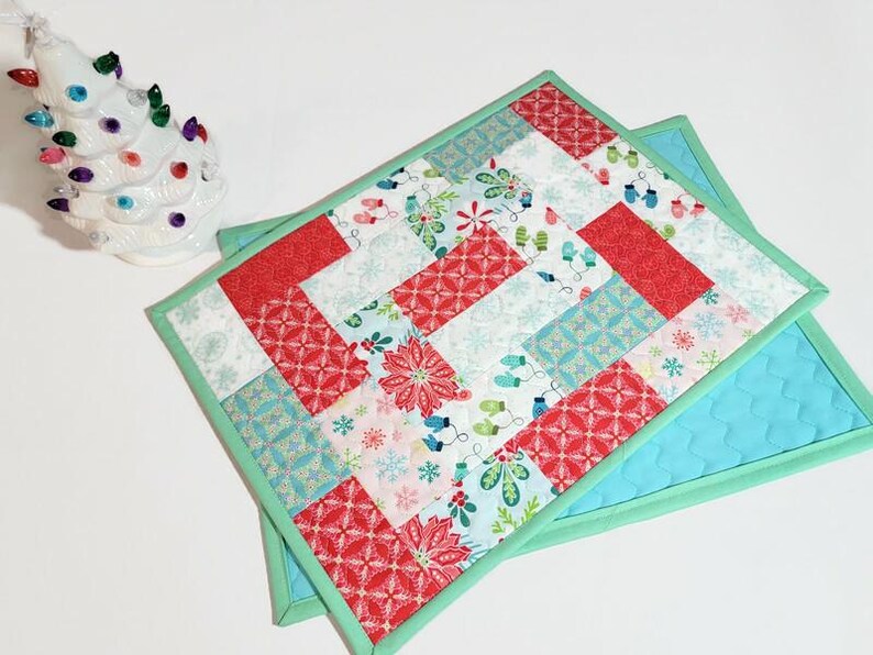 May include: Two quilted placemats with a Christmas theme. The placemats are made of red, white, blue, and green fabric with a variety of patterns. The placemats are edged in a light green fabric.