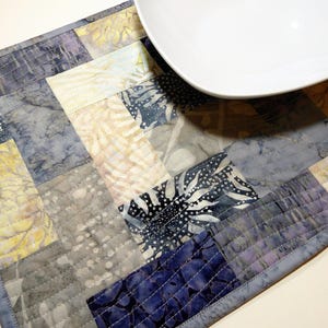 May include: A quilted placemat with a patchwork design in shades of blue, grey, and white. The placemat is made of fabric with a batik pattern and has a stitched border.