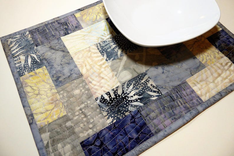 May include: A quilted placemat with a patchwork design in shades of blue, grey, and white. The placemat is made of fabric with a batik pattern and has a stitched border.