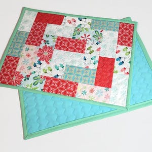 May include: Two quilted placemats with a patchwork design featuring red, green and white holiday-themed fabrics. The placemats have a light blue backing and a green border.