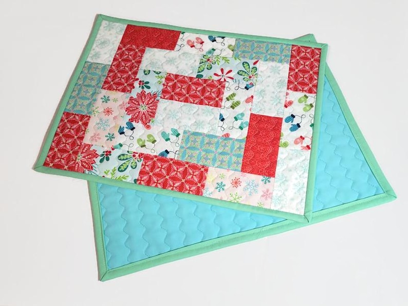 May include: Two quilted placemats with a patchwork design featuring red, green and white holiday-themed fabrics. The placemats have a light blue backing and a green border.