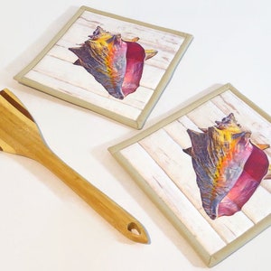 May include: Two beige square fabric pot holders with a brown, yellow, and pink seashell design on a white wood background. A wooden spatula with brown stripes is also in the image.