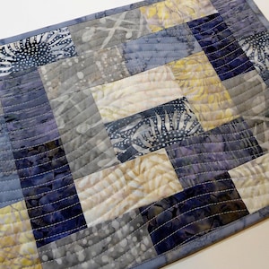 May include: A quilted table runner with a patchwork design featuring various shades of blue, grey, and white. The fabric has a subtle floral pattern.